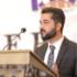 A man in a suit and patterned tie speaks at a podium with a microphone, possibly addressing topics related to Bookkeeping and Accounting Services. The background is blurred with indistinct text and graphics.
