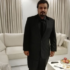 A man with dark hair and a beard, dressed in a black suit, stands in a modern living room with white sofas and beige curtains, ready to discuss Bookkeeping and Accounting Services over food and drinks at the coffee table.
