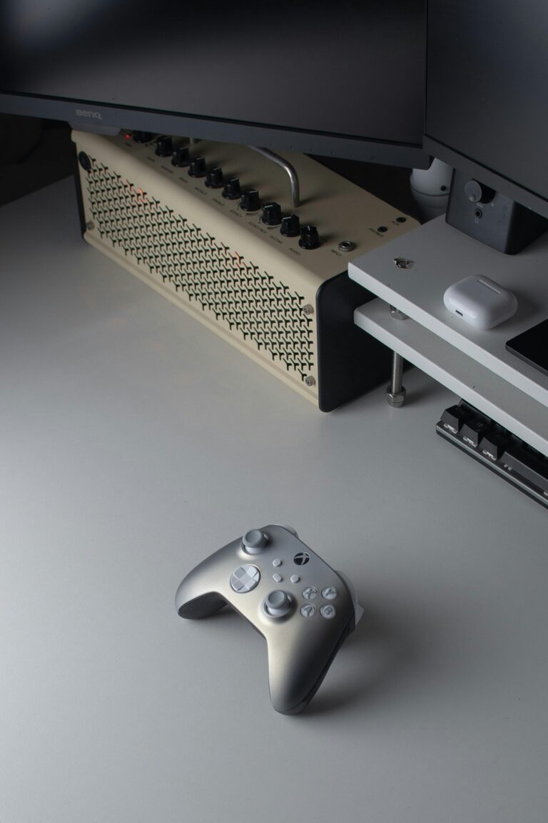 A silver and white game controller rests on a white desk near a monitor, a guitar amplifier, wireless earbuds in a case, part of a keyboard, and materials related to Bookkeeping and Accounting Services.